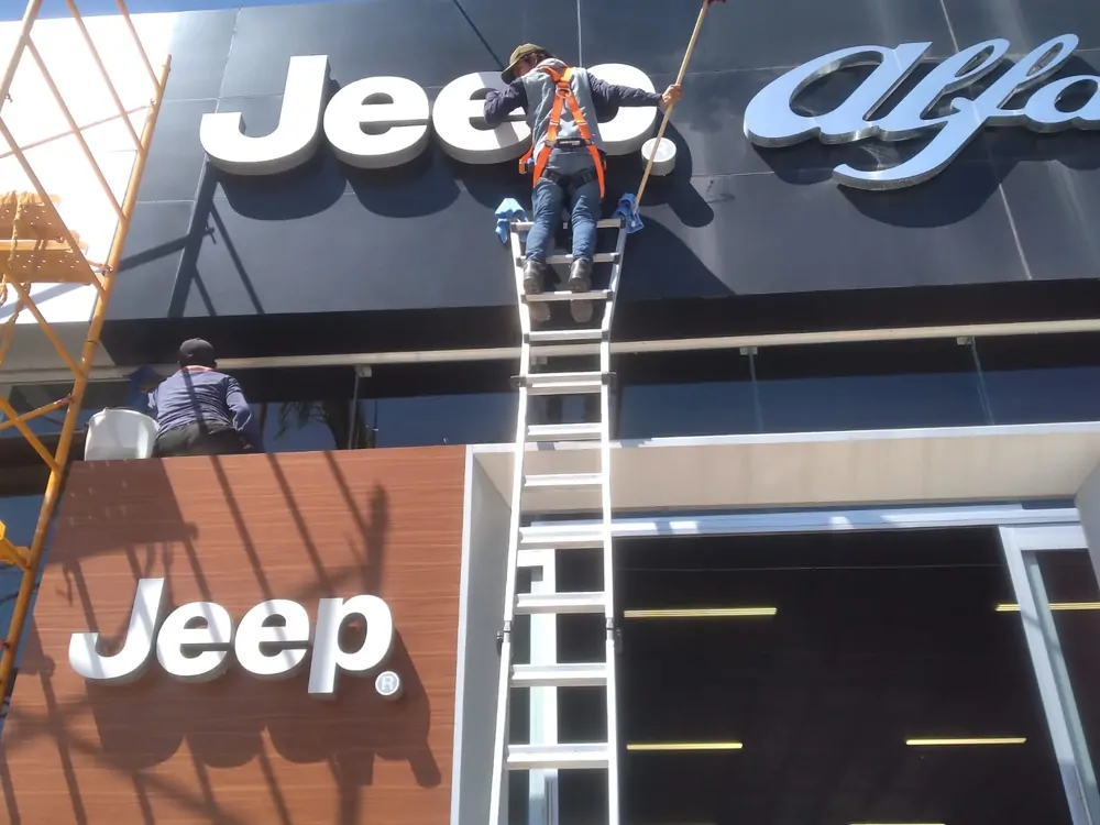 Limpieza de anuncio y fachada de mediana altura en una agencia automotriz, con arnés y escalera